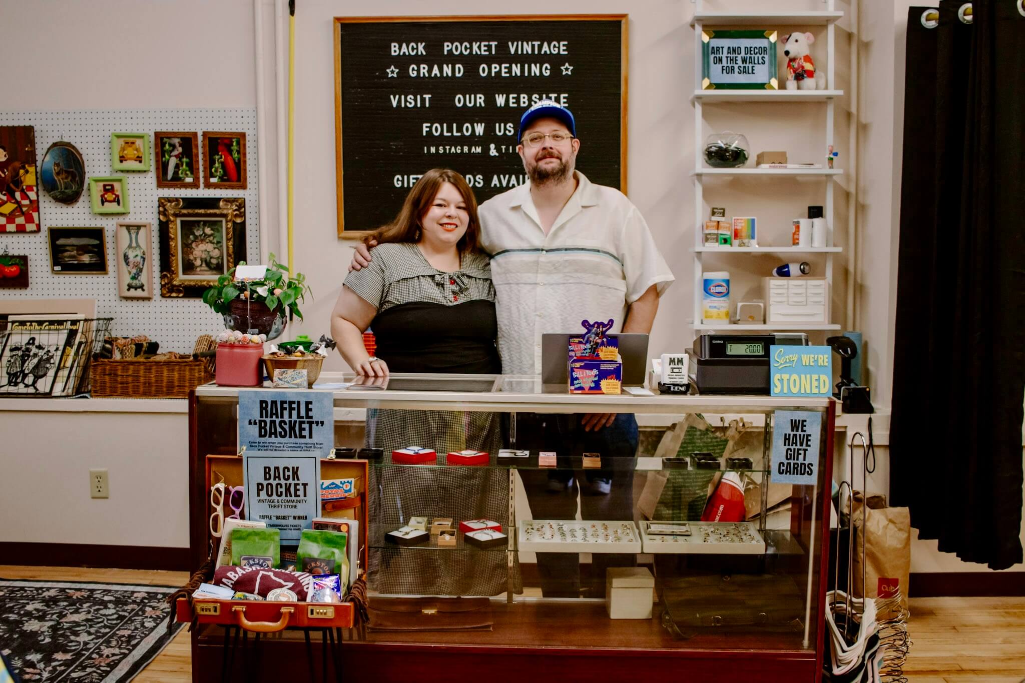 Back Pocket Vintage The owners of Back Pocket Vintage standing behind the shop counter, smiling together in their St. Paul vintage store surrounded by retro décor, framed art, and curated vintage goods.